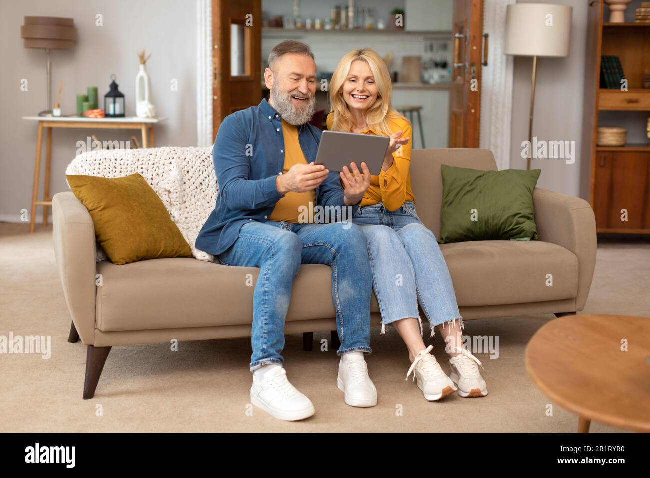 Cheerful Elderly Spouses Using Tablet Sitting On Couch At Home Stock Photo