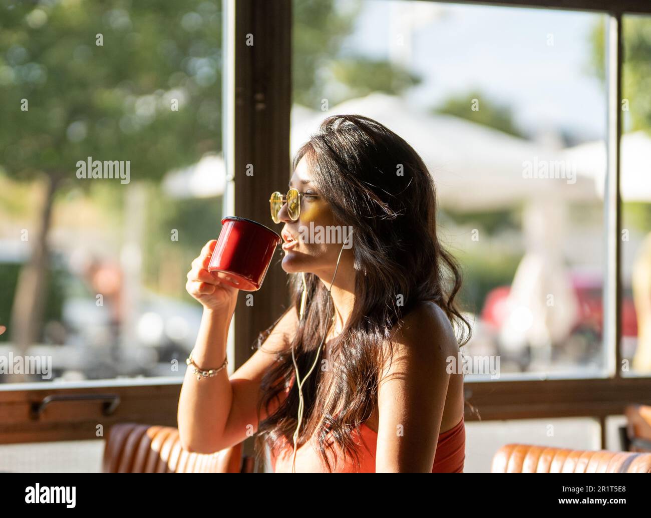 Woman in sunglasses drinking a cup of coffee while relaxing listening music with headphones in a coffee shop. Stock Photo