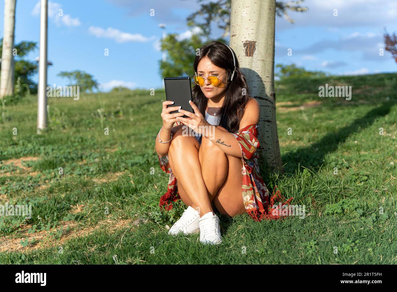 Woman with sunglasses and headphones enjoying reading an ebook while relaxing sitting on grass in a park. Technology concept. Stock Photo