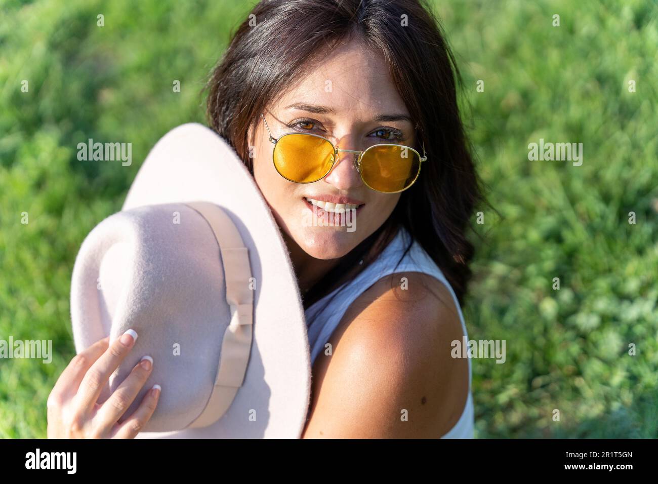 Close-up view of a woman in sunglasses looking at camera and smiling while sitting outdoors on the grass on a sunny day. Stock Photo