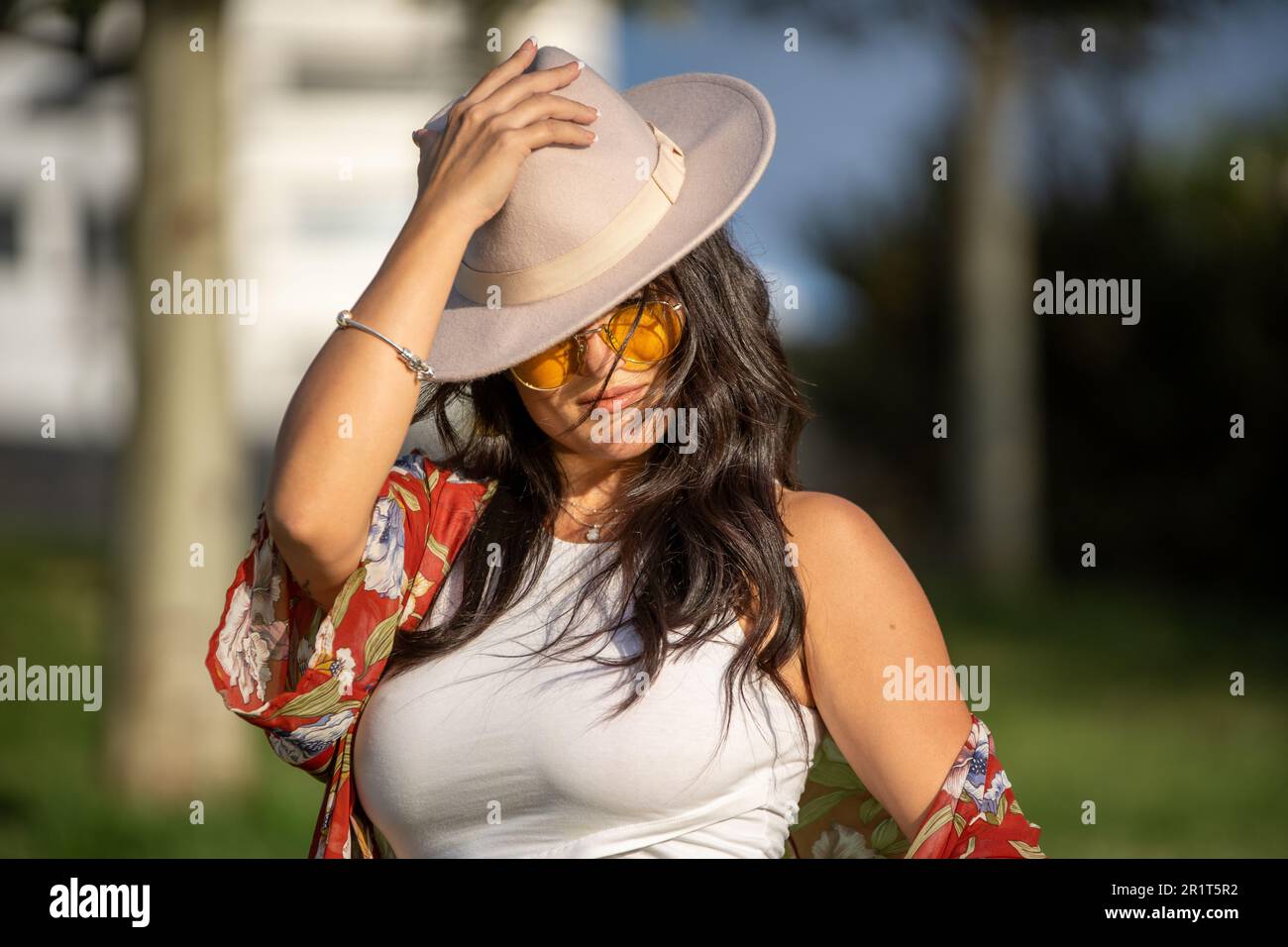 Close-up view of a stylish woman with sunglasses putting on a hat on her head while standing outdoors on a sunny day. Stock Photo