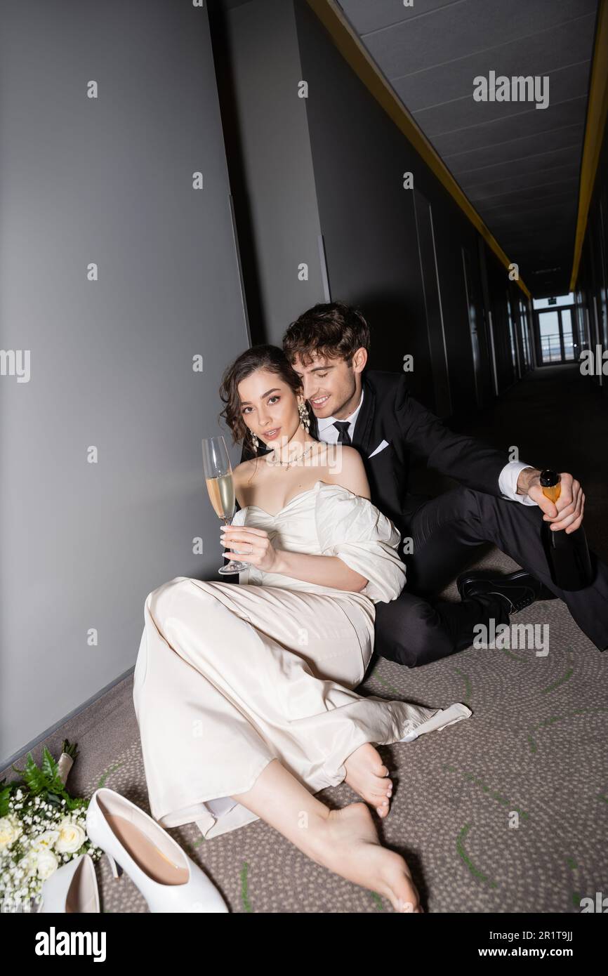 happy groom in black suit holding bottle and sitting near charming bride  with glass of champagne next to bridal bouquet and high heels on floor in  cor Stock Photo - Alamy