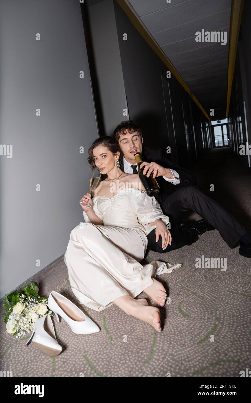 groom in black suit drinking from bottle and sitting near gorgeous bride with glass of champagne next to bridal bouquet and high heels on floor in hal Stock Photo