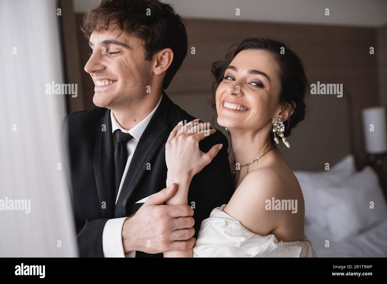 joyful bride in elegant jewelry and wedding dress hugging shoulder of ...