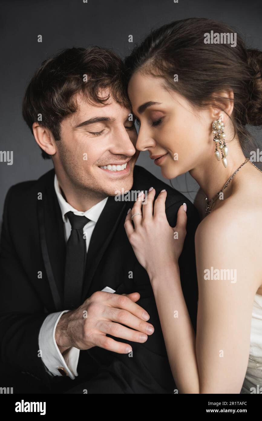 portrait of bride with brunette hair, elegant jewelry and white dress hugging shoulder of happy groom in classic black suit with tie in modern hotel s Stock Photo