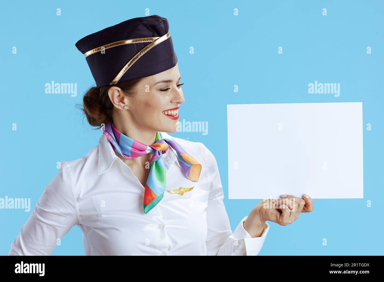 smiling modern female flight attendant isolated on blue background in uniform showing blank a4 paper sheet. Stock Photo
