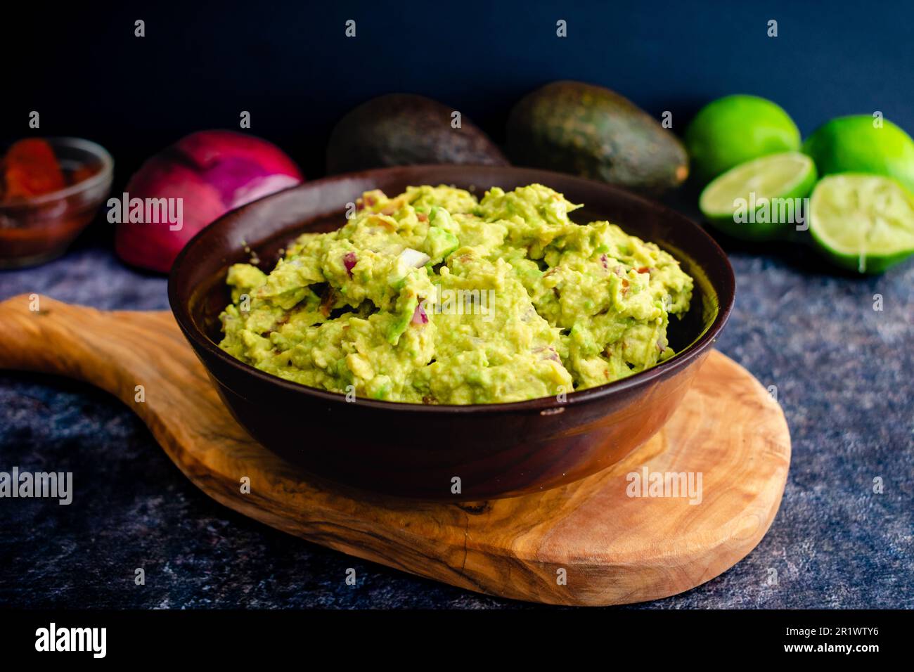 Fresh Guacamole in a Wooden Bowl with Ingredients in the Background: Homemade guacamole with avocados, limes, onion, cilantro, and chipotle peppers Stock Photo