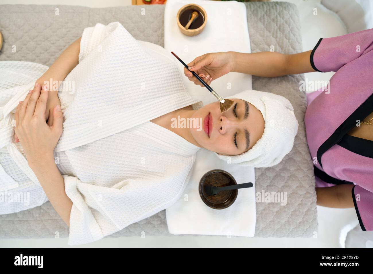 Young asian woman with nutrient facial dressed in bathrobe, getting facial care by beautician at