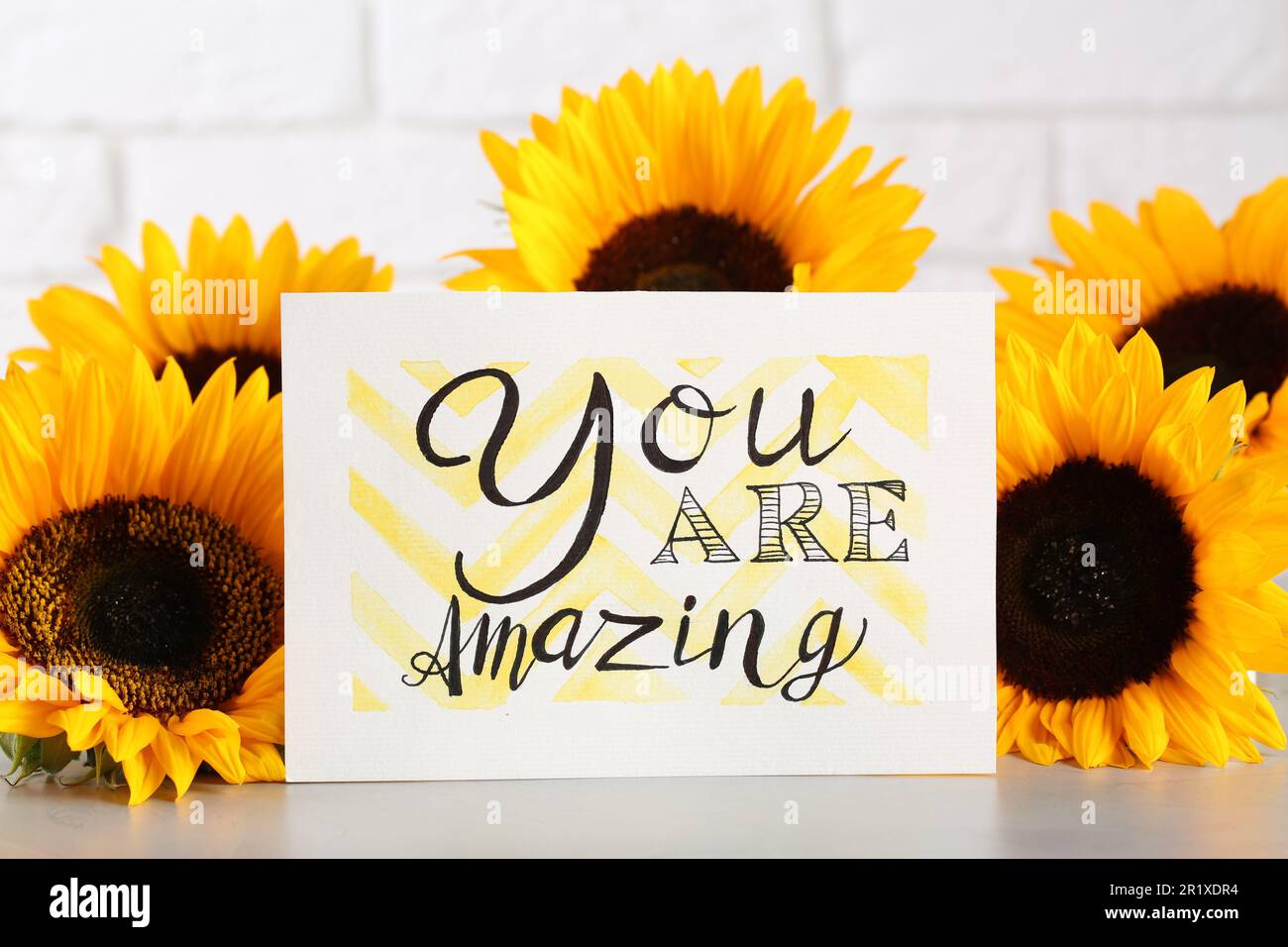 Card with life-affirming phrase You Are Amazing and sunflowers on light table against white brick wall Stock Photo