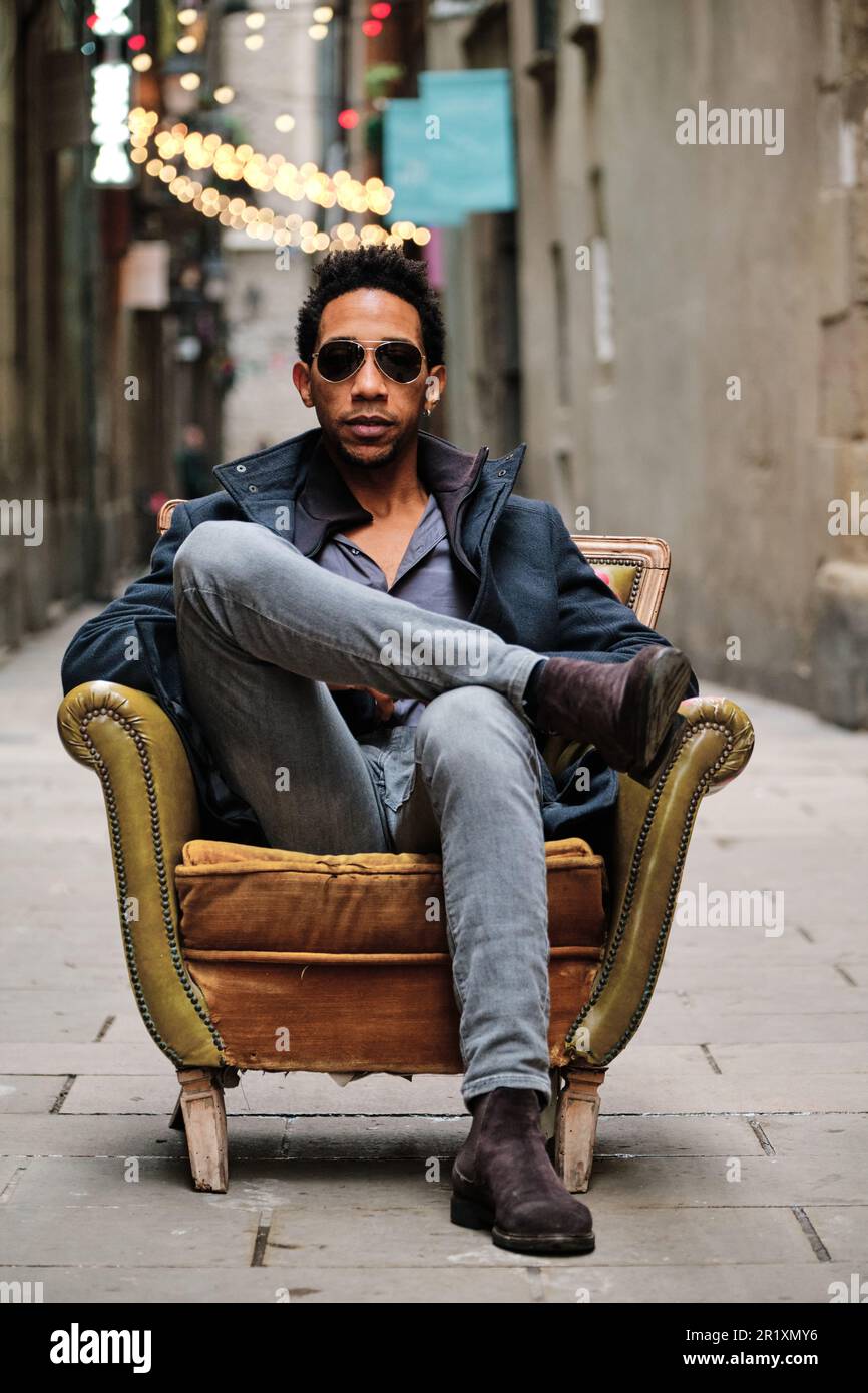 Serious man with sunglasses looking at camera while sitting on an armchair outdoors. Stock Photo