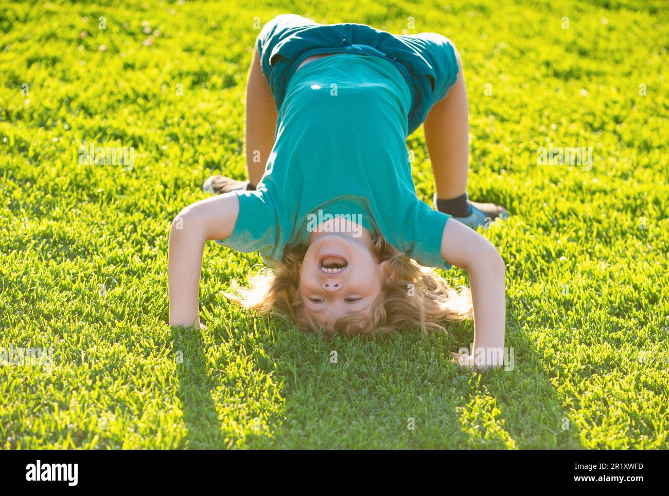 Happy kid boy girl standing upside down on her head on grass in summer