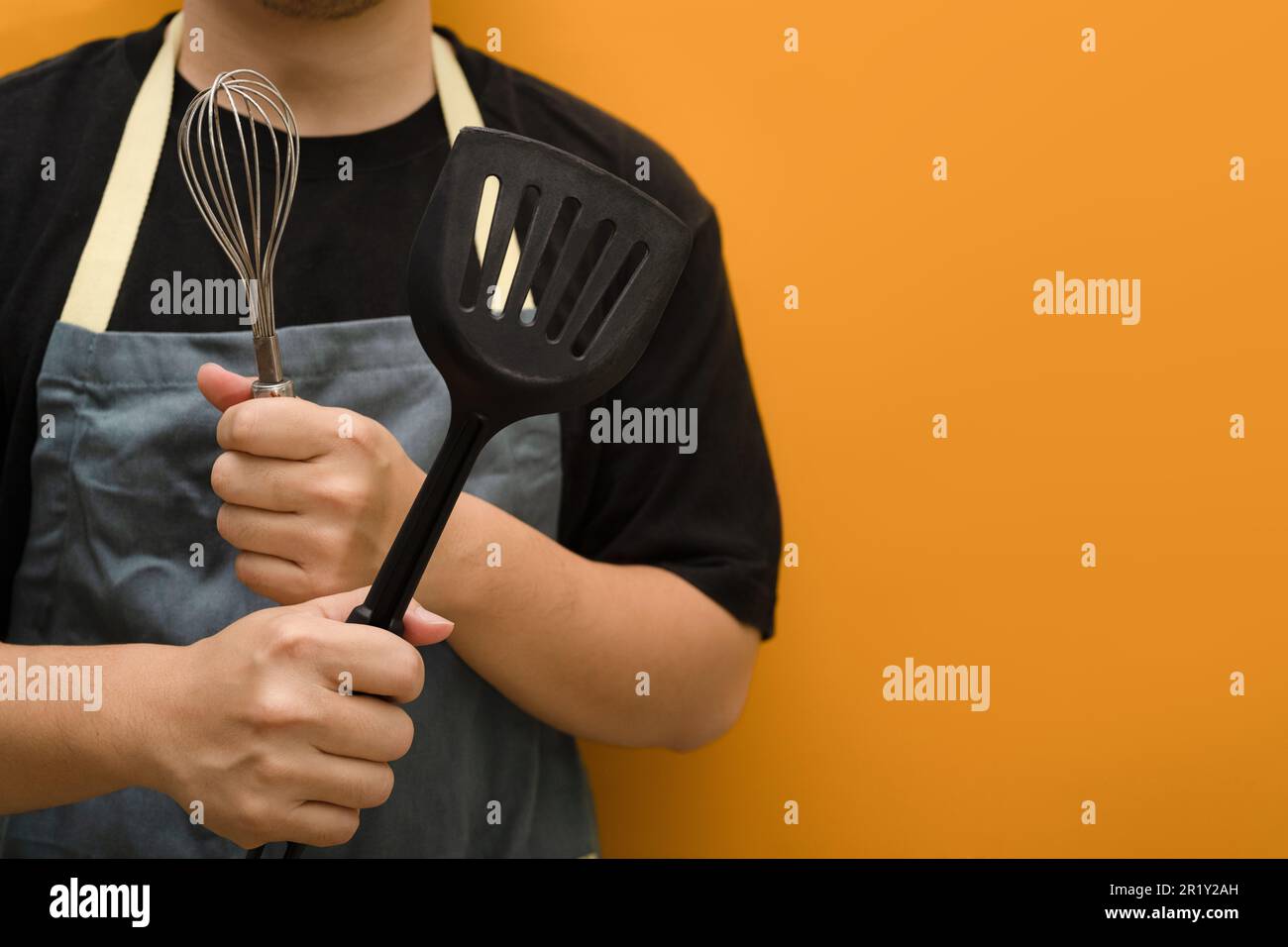 Cooking food concept. Man in apron holding whisks and kitchen spatula standing against yellow background with copy space Stock Photo
