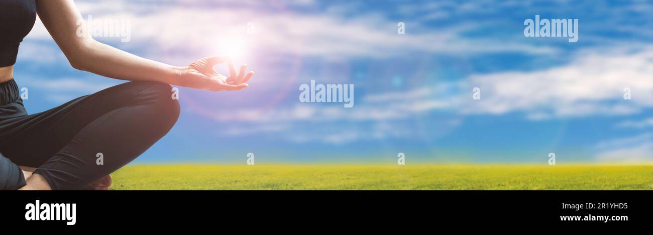 Yoga meditation zen-like, woman meditating on background of blue sky and green meadow holding lens flare in hand. Stock Photo