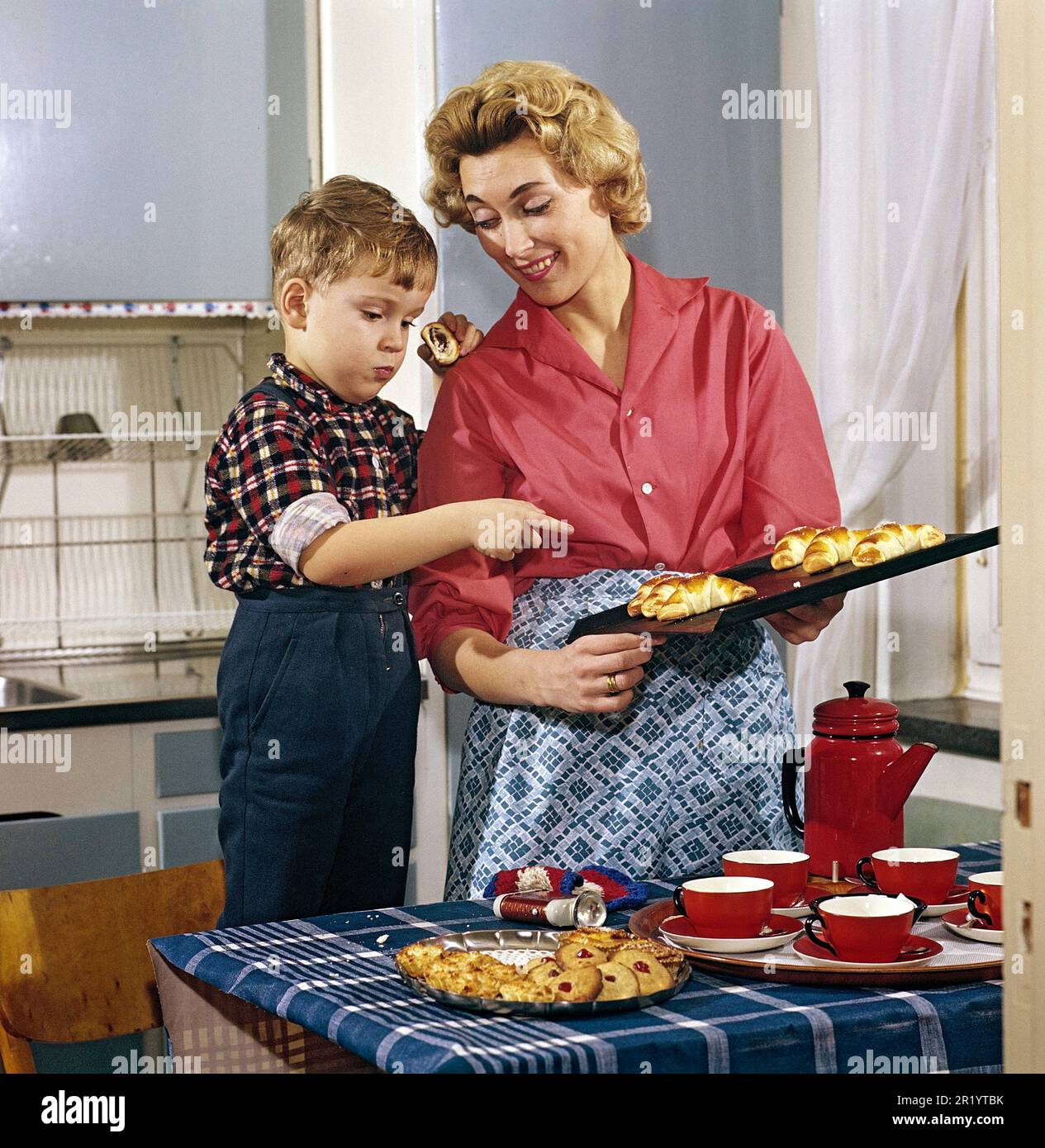 Baking in the 1950s. A mother with her son in the kitchen. She has freshly baked buns and cakes from the oven and the boy looks as if he likes the nice smell from them. A typical 1950s picture with the mothers clothes, the cups and can, the kitchen interior and the colors. Sweden 1958 Conard Stock Photo