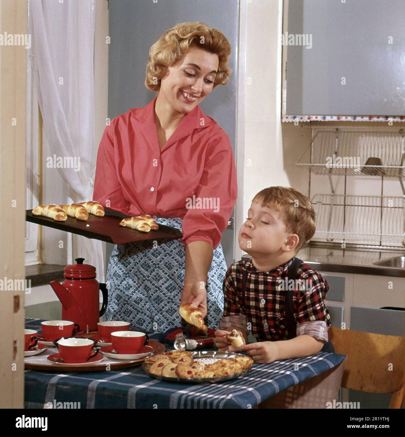 Baking in the 1950s. A mother with her son in the kitchen. She has freshly baked buns and cakes from the oven and the boy looks as if he likes the nice smell from them. A typical 1950s picture with the mothers clothes, the cups and can, the kitchen interior and the colors. Sweden 1958 Conard Stock Photo