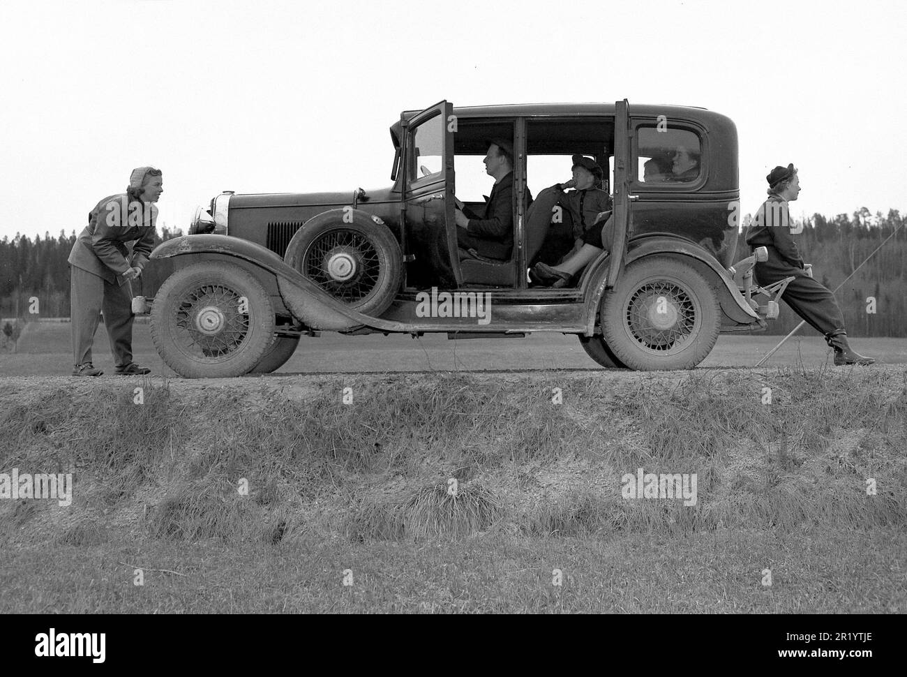 The old car. A family out driving with their vintage car looks as if it's stopped and they are trying to start it again. The early cars could also be started with a crank, if the battery had ran out of power. A woman is seen using it in front of the car while the man is behind the wheel. By the look of the passangers faces and body language this may have been a long ongoing attempt.   Sweden 1952. Kristoffersson ref 12K-24 Stock Photo