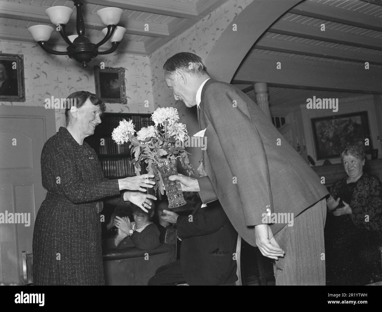 Birthday in the 1930s. An elderly lady recieves a flowers in a vase with peonies flowers by an equally old gentlemen, bowing politely when handing them over. People in the room behind them are applauding. They are residents of the retirement home Höstsol where older actors live in their golden days. November 1939. Kristoffersson ref 40-4 Stock Photo