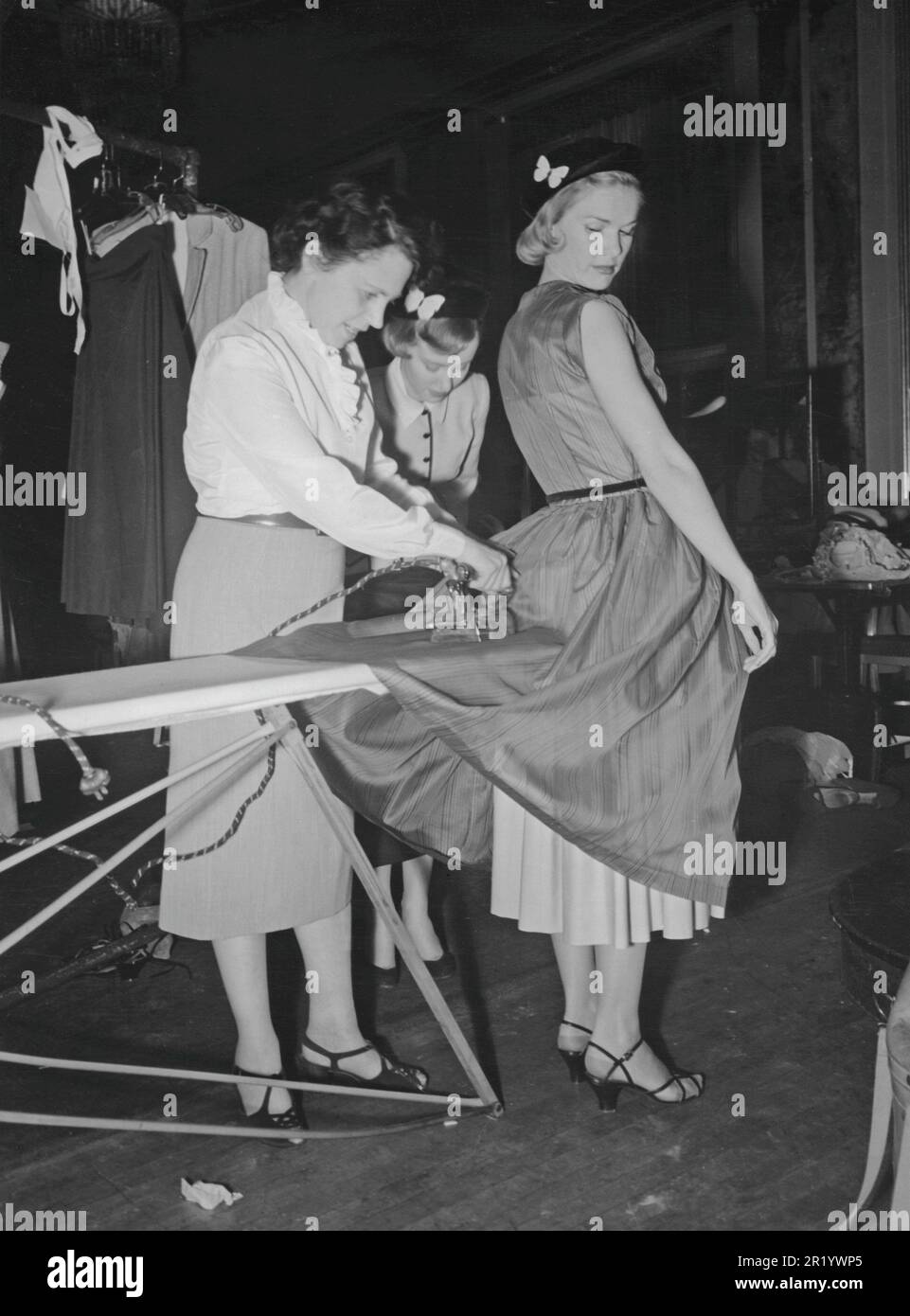 Women's fashion in the 1950s. The young fashion model stands absolutely still while an assistant irons her dress just moments before the model goes on stage to show it. It's in the details of course and a behind the scenes photograph. Sweden 1951 Stock Photo