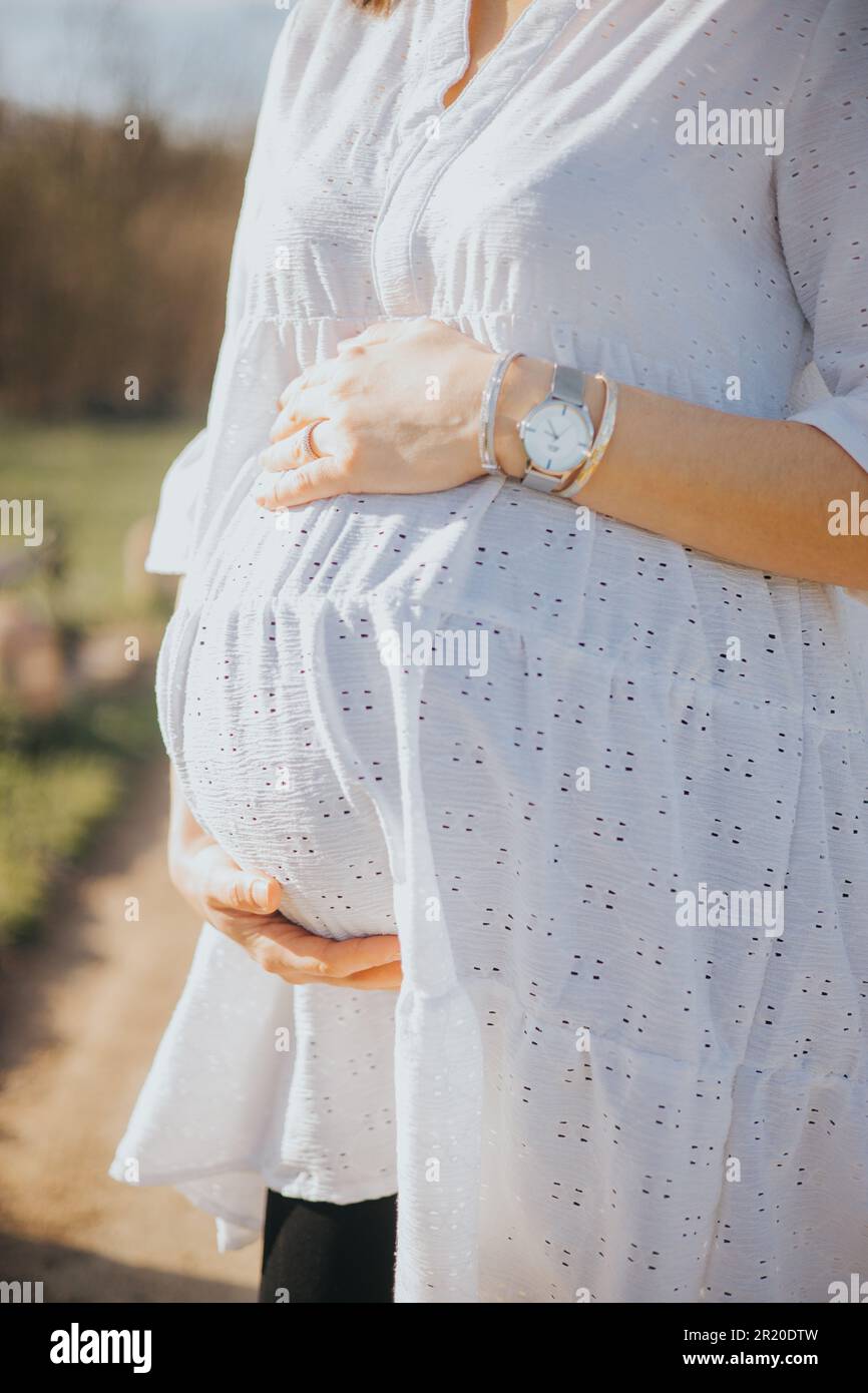 A young pregnant woman with a white dress holding her belly in a park ...