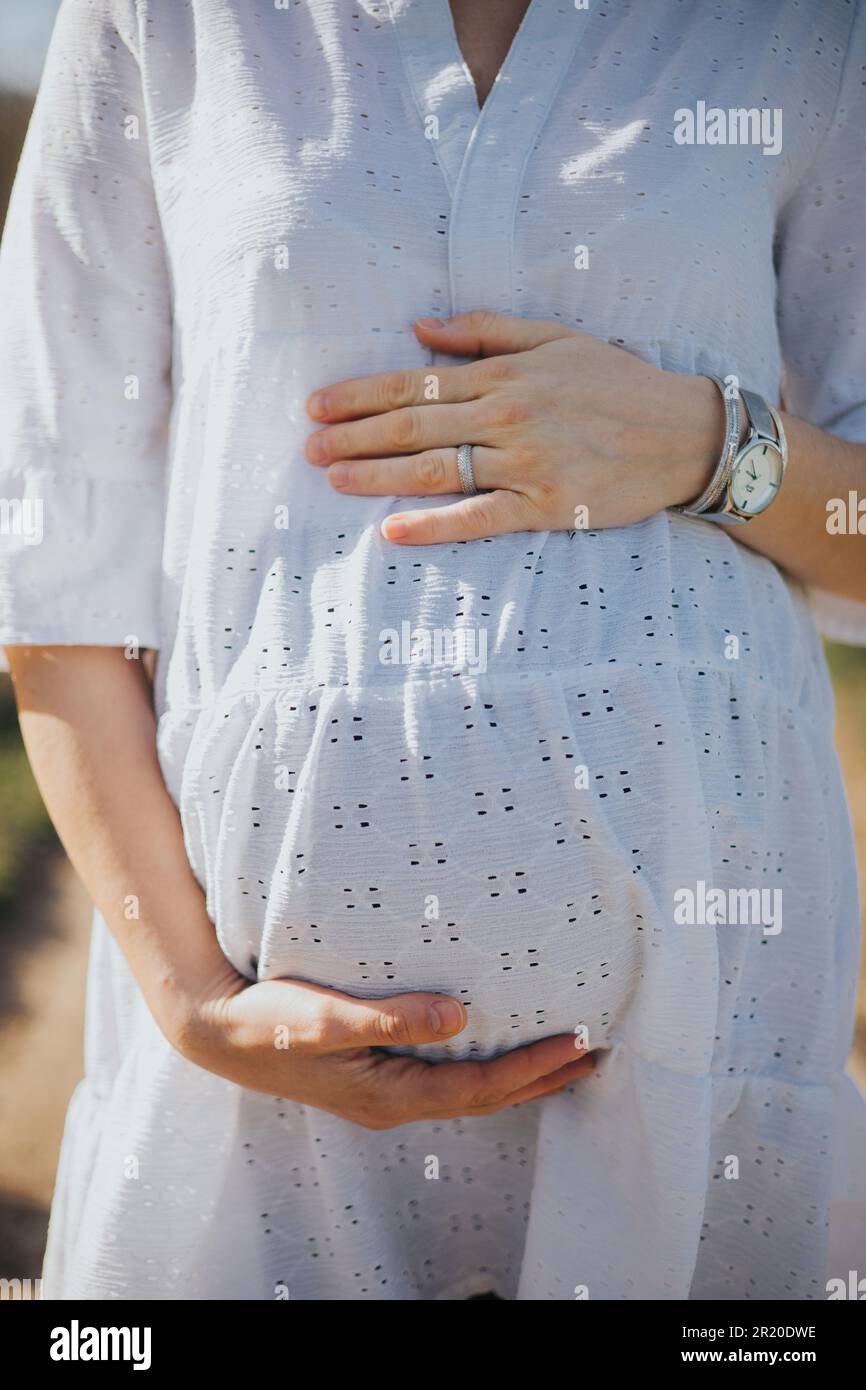 A young pregnant woman with a white dress holding her belly in a park ...