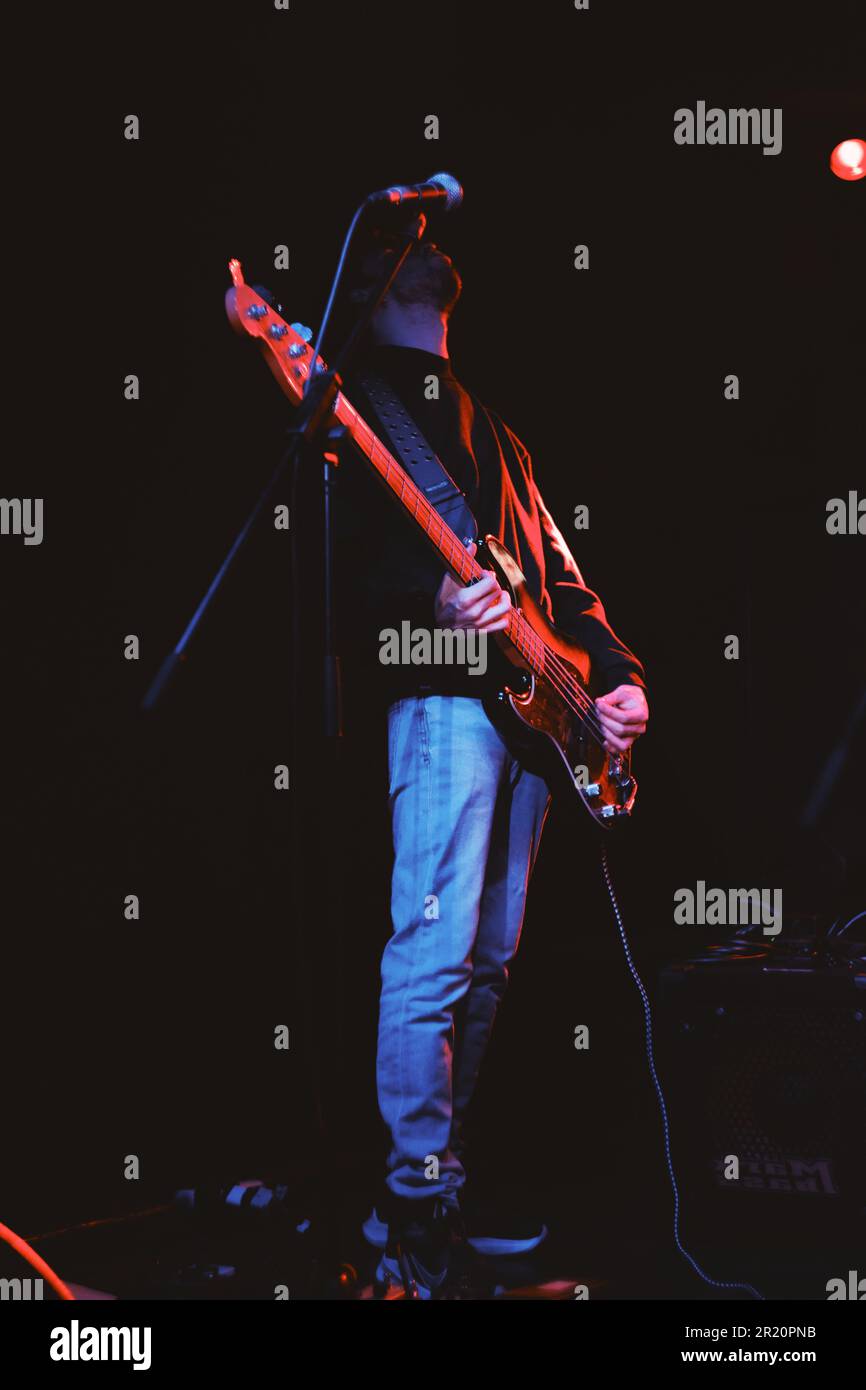 A confident young man strums an electric guitar on stage in front of a vibrant crowd, illuminated by bright stage lights in a night time setting Stock Photo