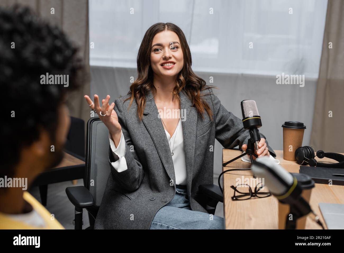 pleased brunette podcaster in grey blazer holding microphone near headphones, notebook, paper cup and eyeglasses while gesturing and talking to indian Stock Photo