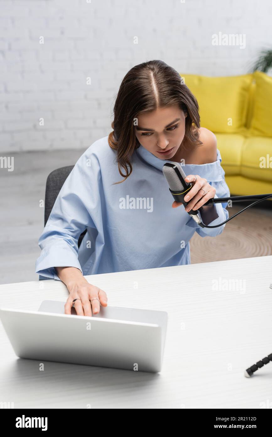 Brunette broadcaster in blue blouse talking at microphone and using blurred laptop while sitting near table during online stream in podcast studio wit Stock Photo