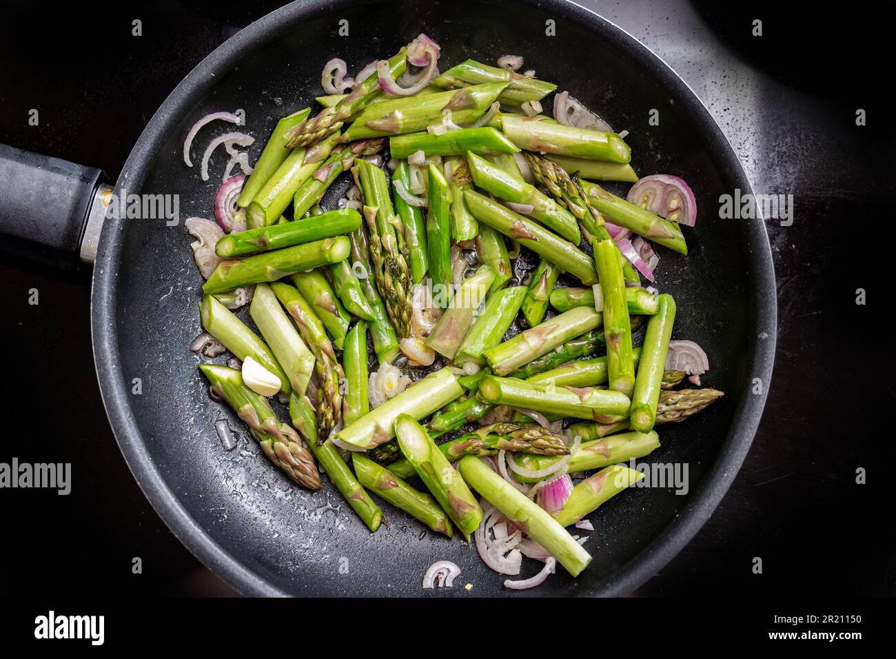 Green asparagus and red onion in a frying pan on the black stove top, cooking a healthy vegetarian meal with seasonal vegetables, high angle view from Stock Photo