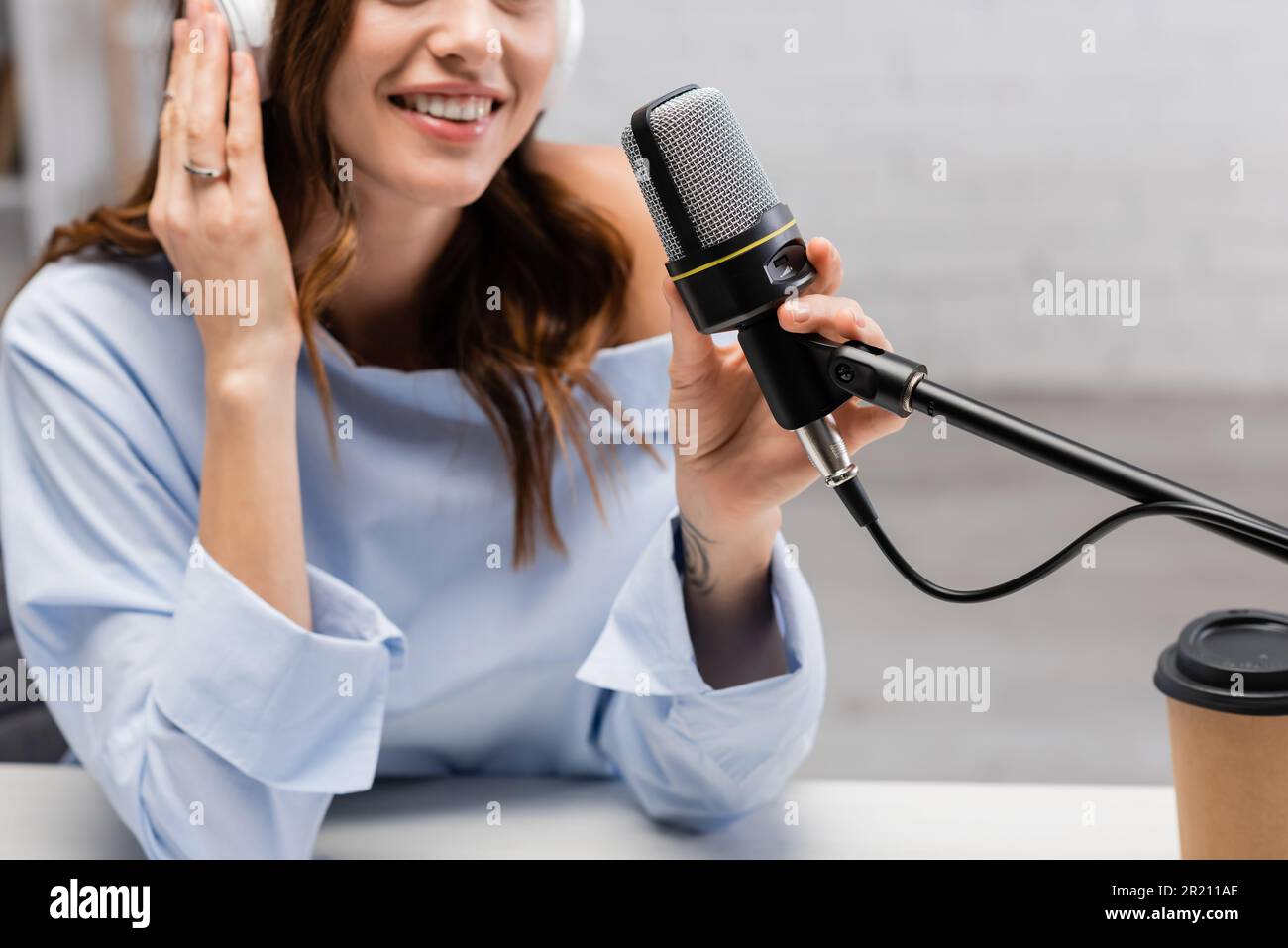 Cropped view of blurred and positive podcast host in wireless headphones talking near microphone and takeaway coffee in paper cup on table in studio,s Stock Photo