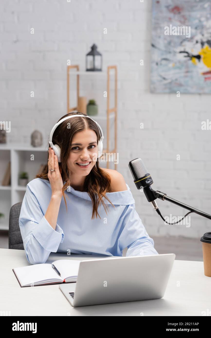 Cheerful brunette podcaster in headphones looking at camera near devices, notebook, paper cup with coffee to go and microphone during stream in podcas Stock Photo