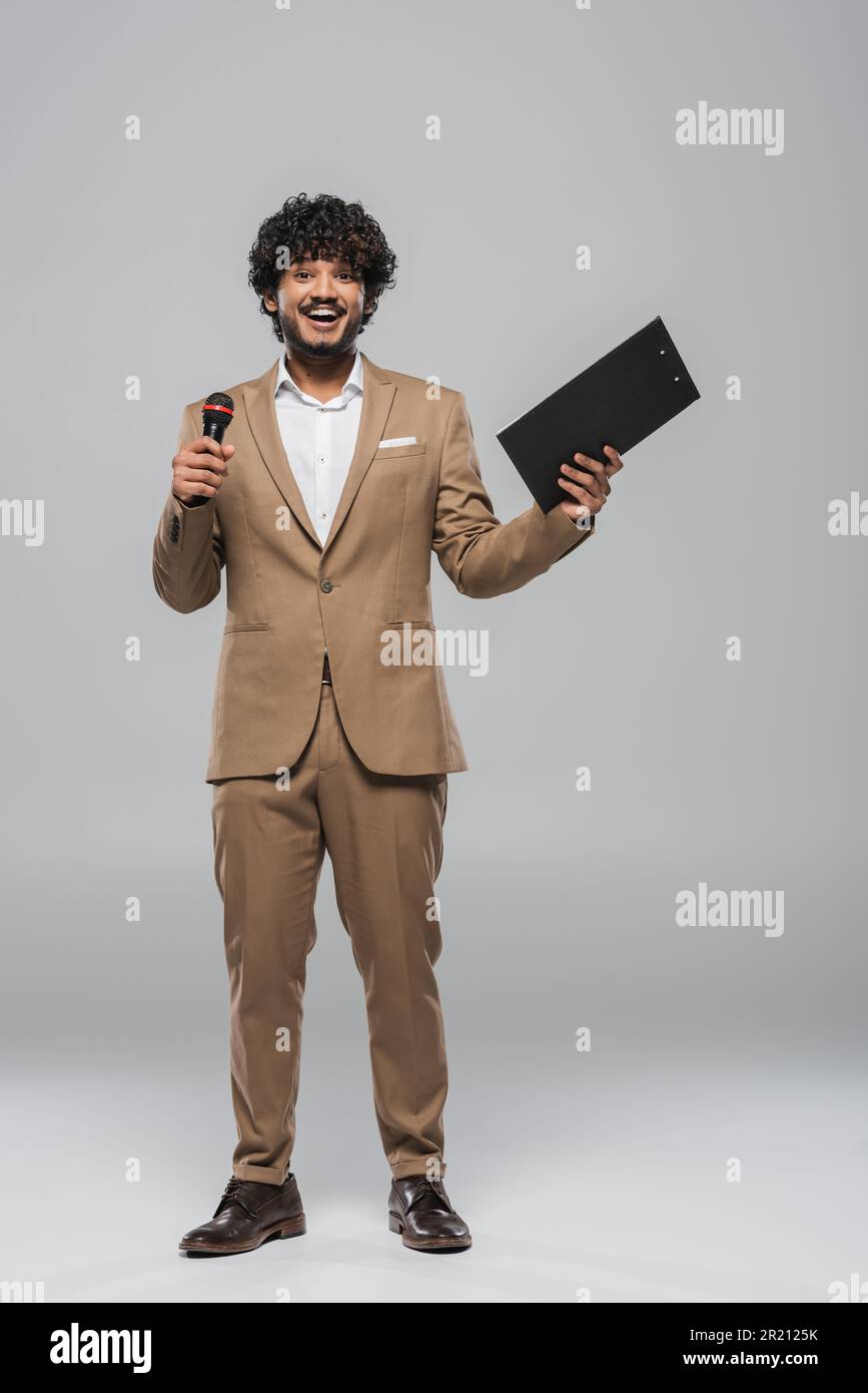 Full length of smiling indian event host in brown suit holding wireless microphone and clipboard during holiday while standing on grey background,stoc Stock Photo