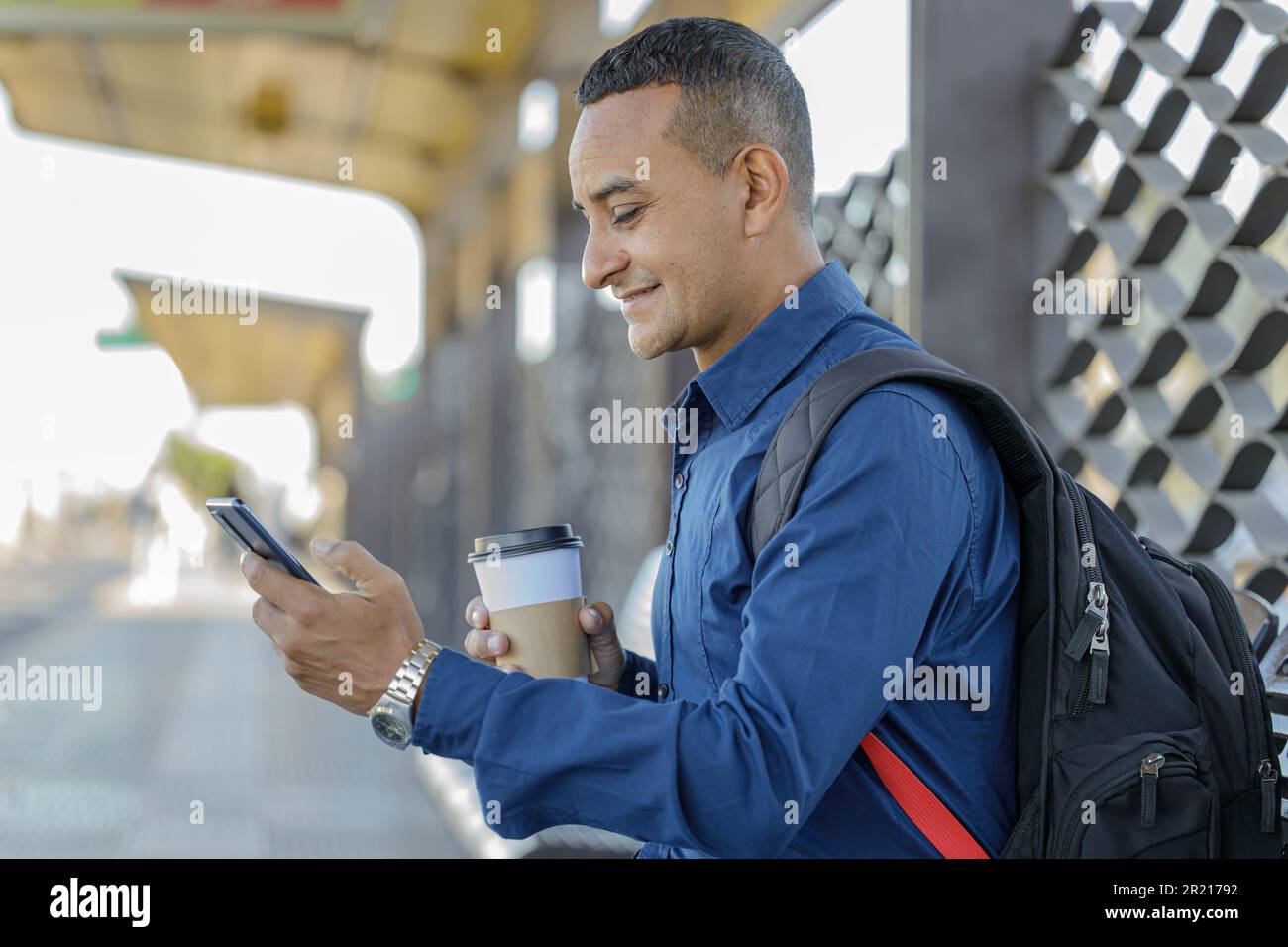 Young latin man looking at his mobile phone with paper cup with coffee in his hand. Stock Photo