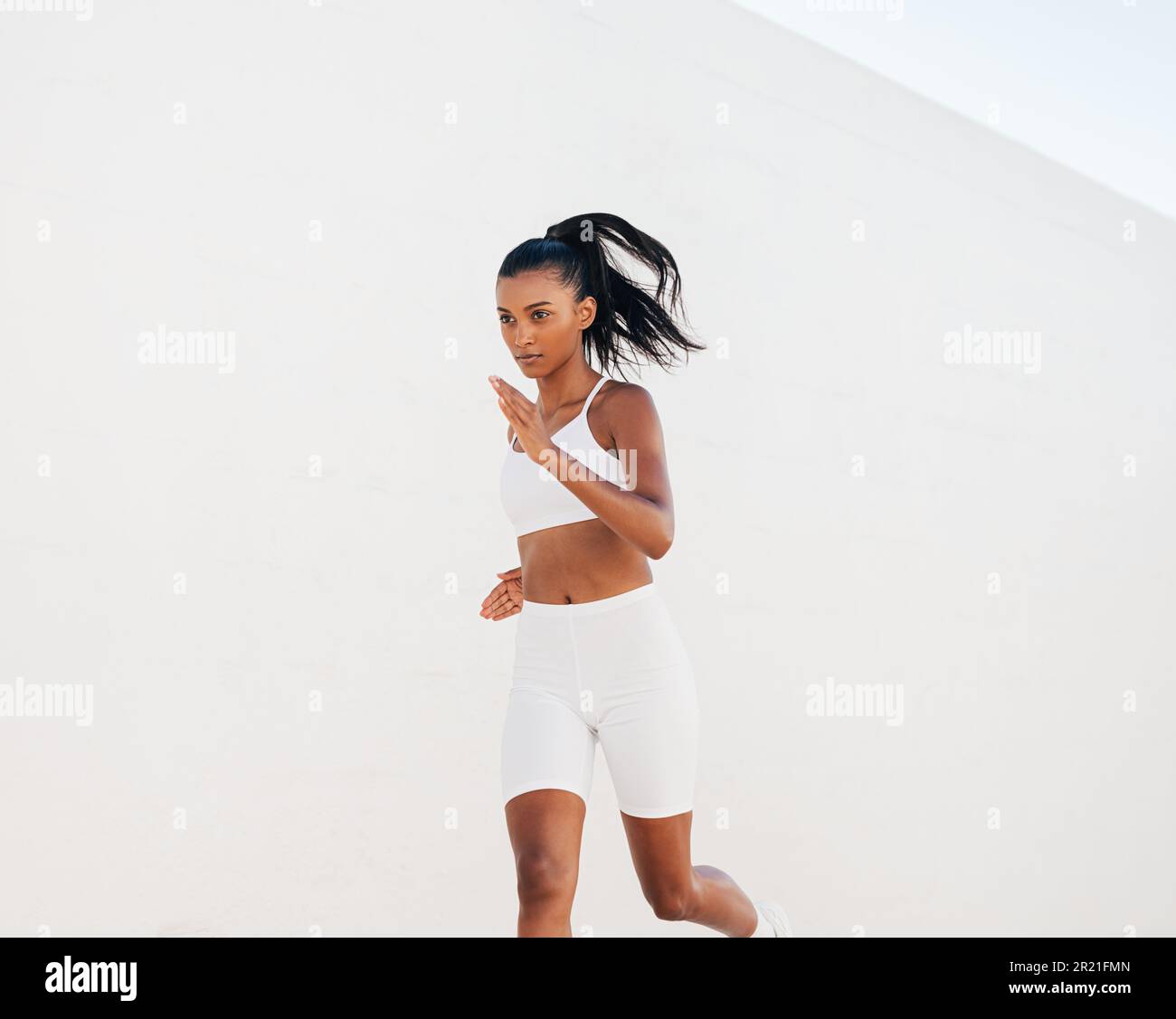 Confident woman sprinting against a white wall. Slim jogger in white fitness attire running outdoors. Stock Photo