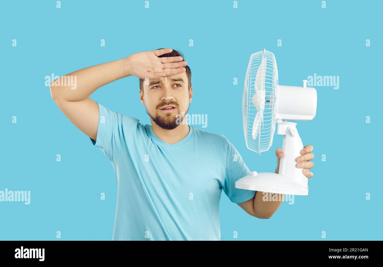 Young emaciated Caucasian man suffers from summer heat keeps fan and wipes sweat with hand Stock Photo
