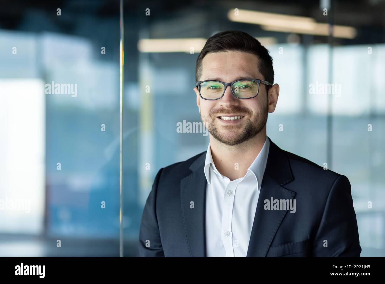 Close-up photo. Portrait of a young businessman, founder, director in a suit and glasses standing in the office and smiling at the camera. Stock Photo