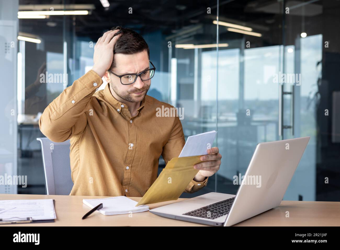Shocked young male businessman sitting in the office at the desk and reading a letter. I received bad news, financial problems, a business deal, bankr Stock Photo
