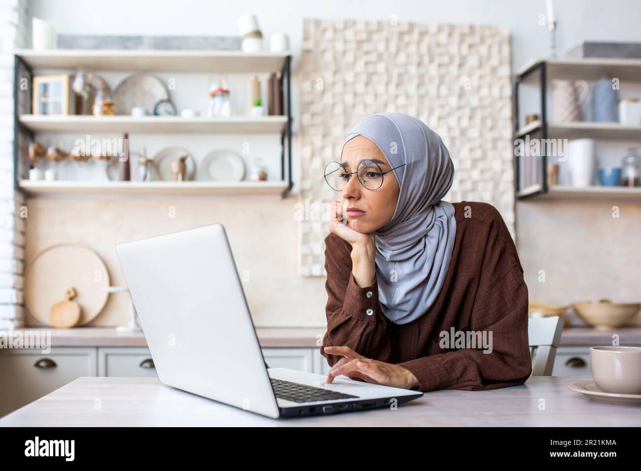 Bored young muslim woman wearing hijab sitting in the kitchen at home ...