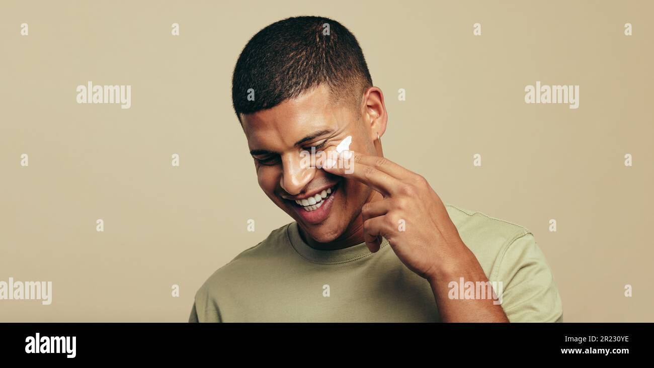 Young man enjoying his grooming and skincare routine as he uses a beauty product on his face. Handsome man applying facial cream with a glowing smile, Stock Photo