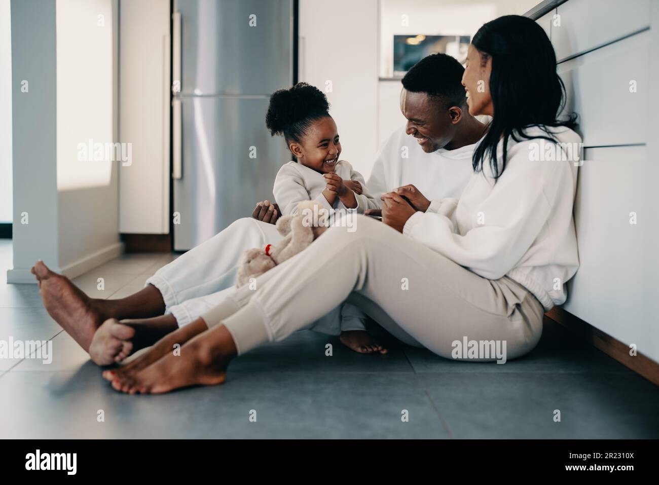 Couple bonding with their child at home. Girl parents playing with their daughter while sitting on the kitchen floor. Little girl enjoying the love an Stock Photo