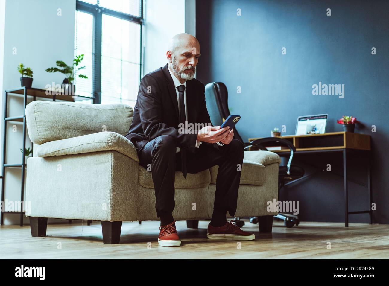 Old businessman dressed in black suit can be seen either texting on phone or browsing internet. Mature man, with distinguished silver beard, sits comfortably in chair within stylish loft office. . High quality photo Stock Photo