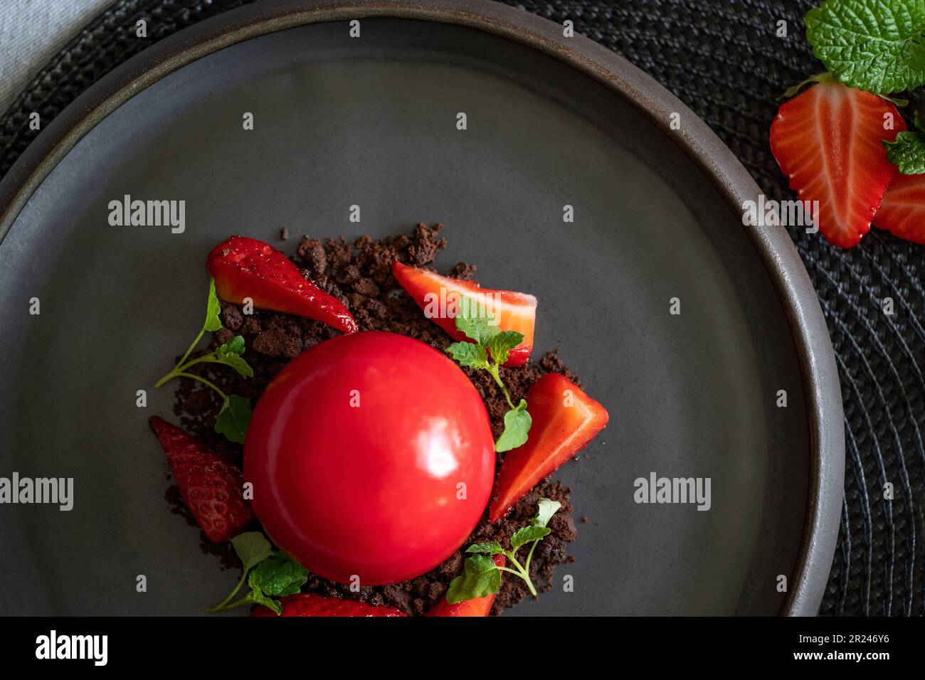 Selective focus of strawberries on the brownie crumbs. Sphere curd cake with smooth surfaces and mirror glaze. Red dessert on the black plate. Stock Photo