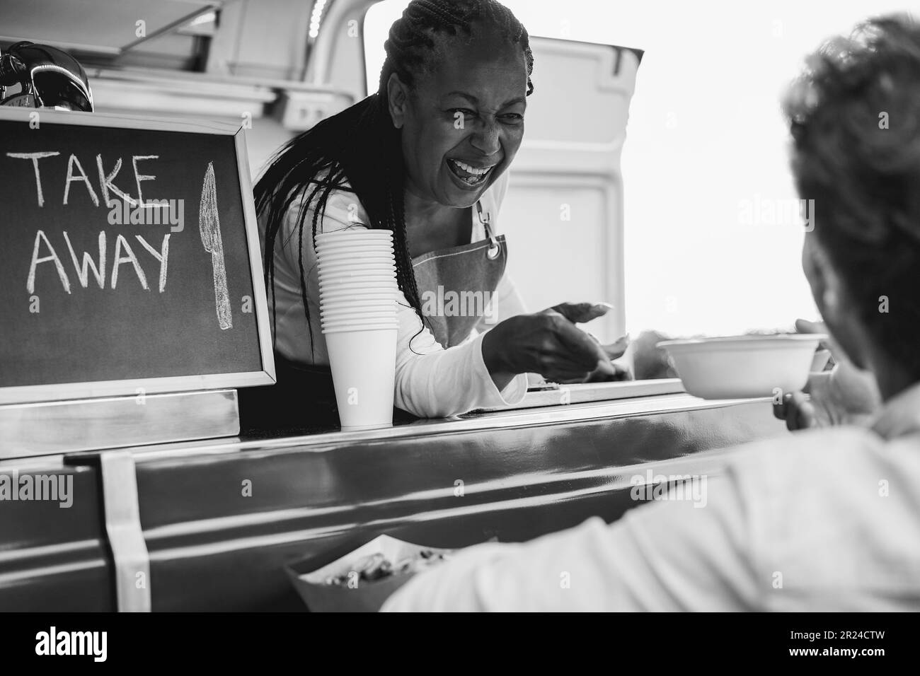 African senior woman serving take away food inside food truck - Soft focus on chef face - Black and white editing Stock Photo