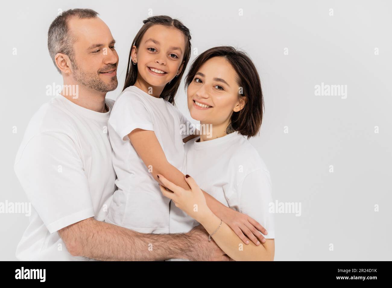 portrait of happy family in white t-shirts looking at camera and hugging each other on grey background, International child protection day, parents an Stock Photo