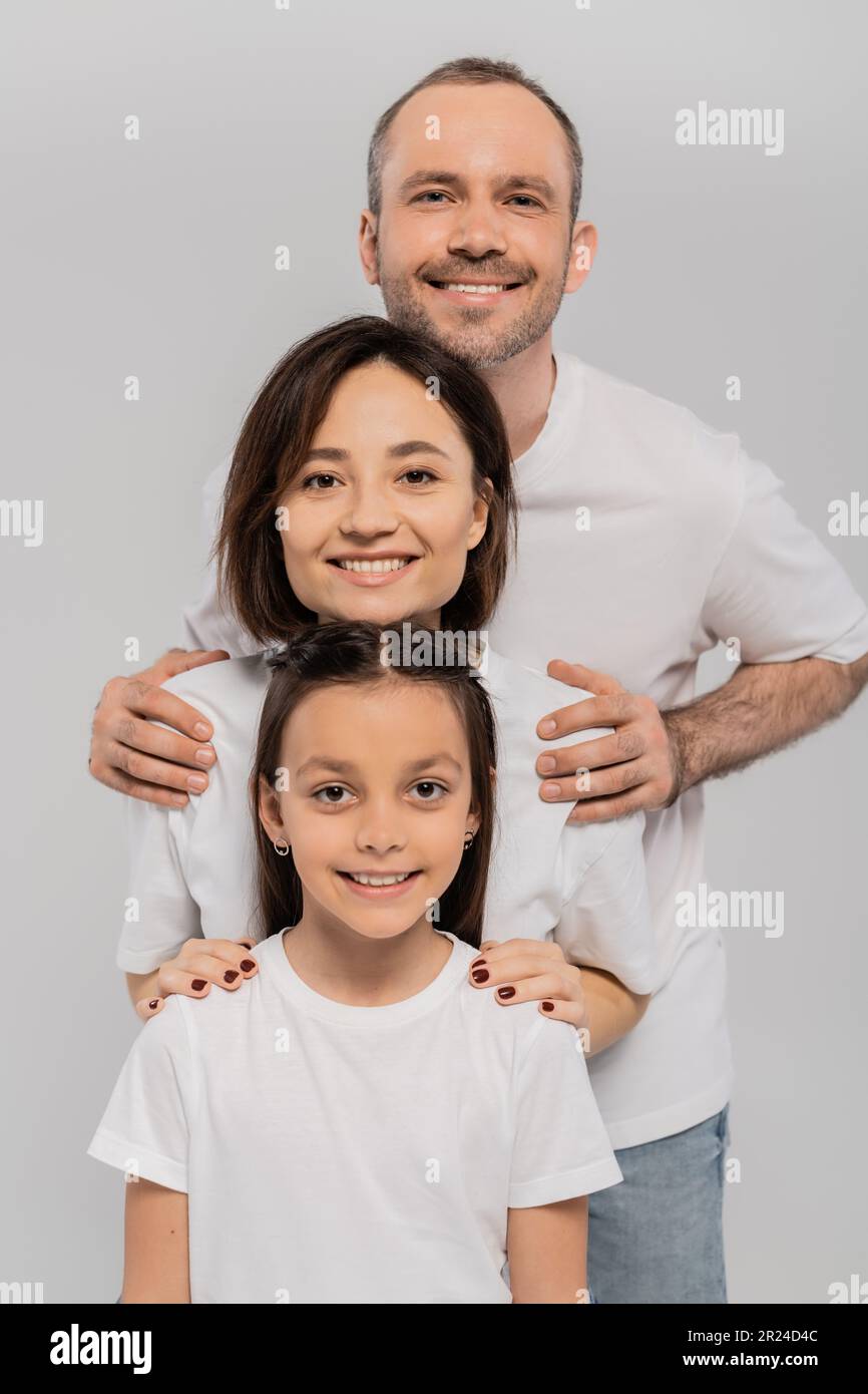 happy family in white t-shirts looking at camera and posing together on grey background, International child protection day, positive parents and daug Stock Photo