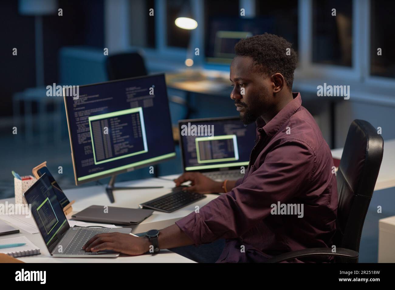 African American developer working with computer program on computer while sitting at his workplace in late evening Stock Photo