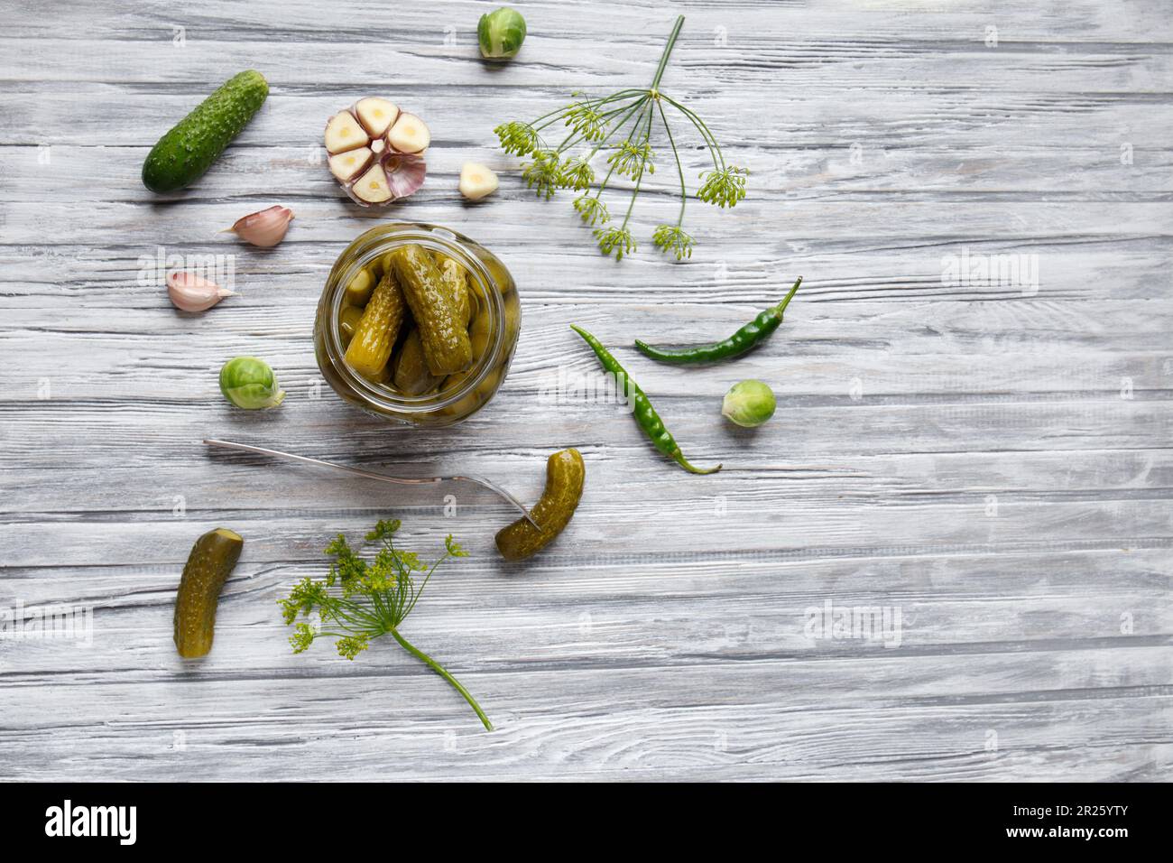 Pickled cucumbers in an open jar, gherkin on a fork and garlic on a light wooden background, top view, copy space. Homemade canned cucumbers. Stock Photo