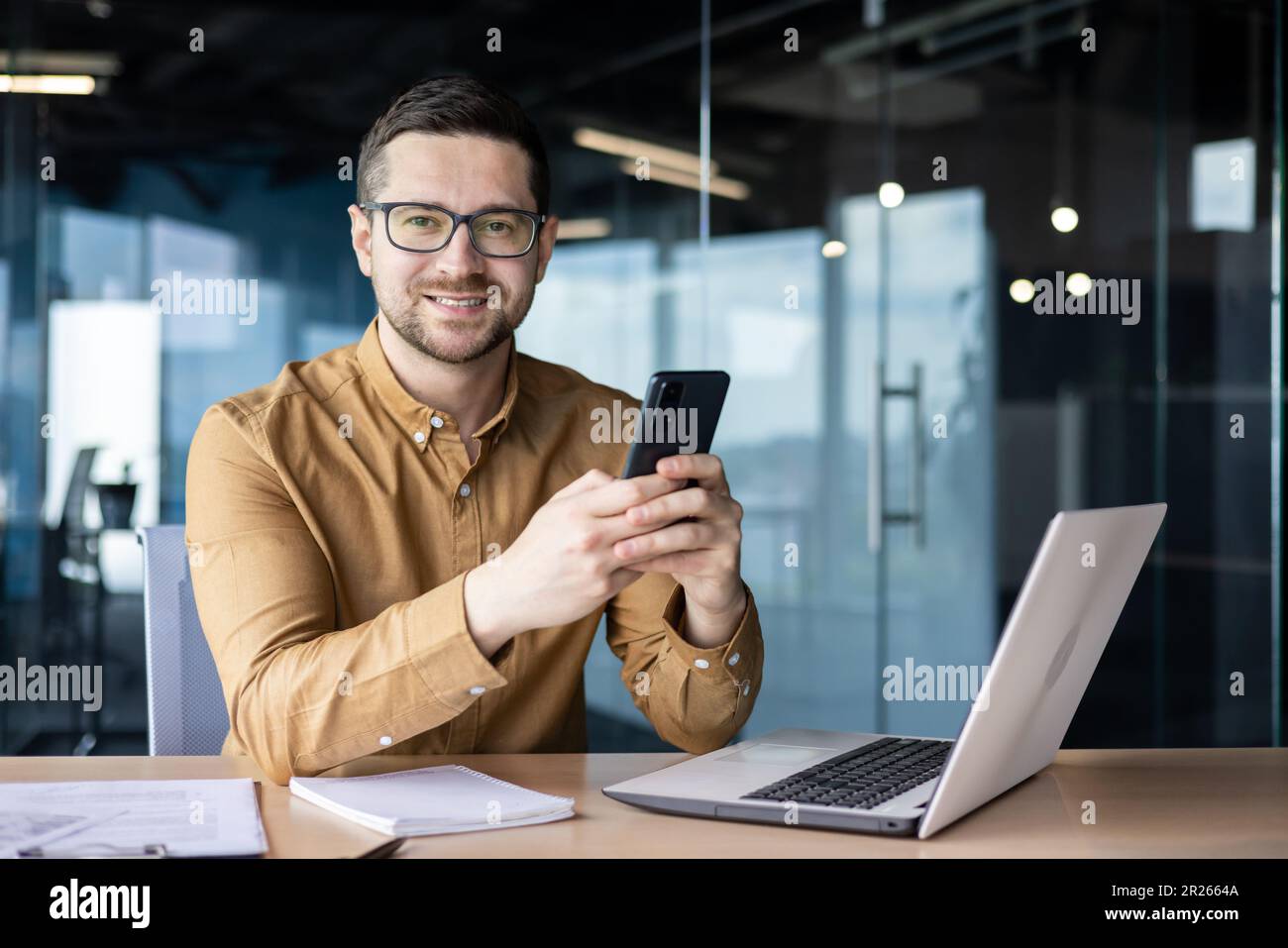 Portrait of a young businessman working in an office center, sitting at a desk and using a mobile phone. Smiling looking at the camera. Stock Photo