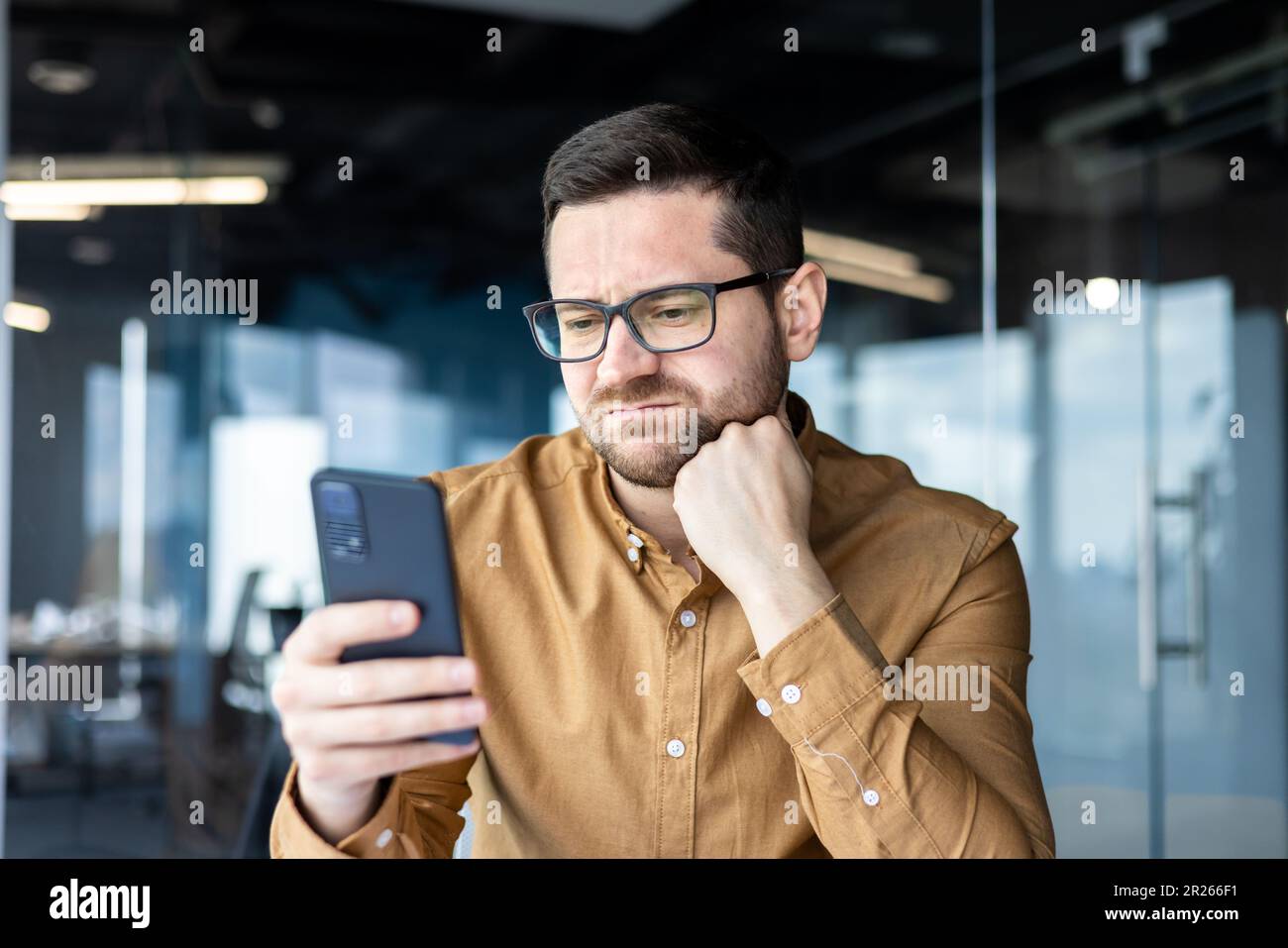 Tired businessman, employee sitting at a desk in an office center and using the phone. Bored and grimacing, he looks at the screen, checks his mail, p Stock Photo
