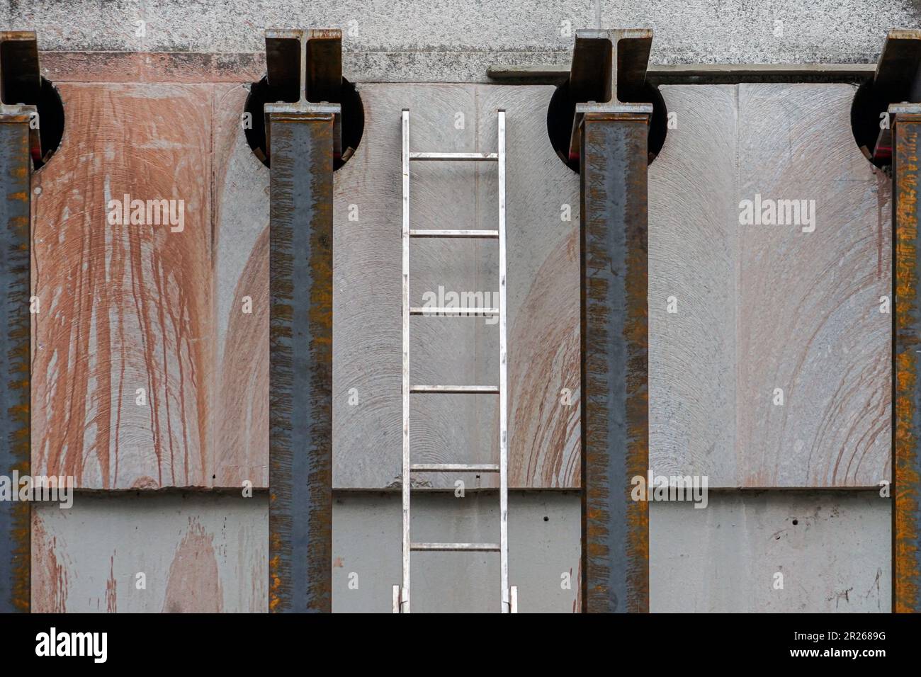 Between two rusty metal columns, an aluminum ladder is leaning against a house wall. Stock Photo