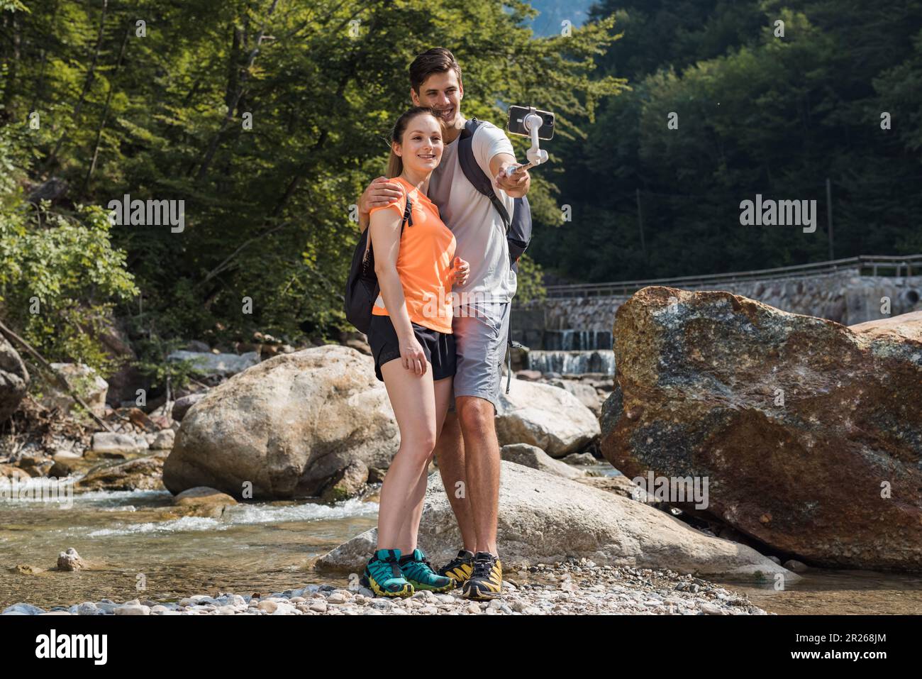 Handsome young couple standing in nature near the mountain river, making selfie vlog videos using a smartphone and selfie stick Stock Photo